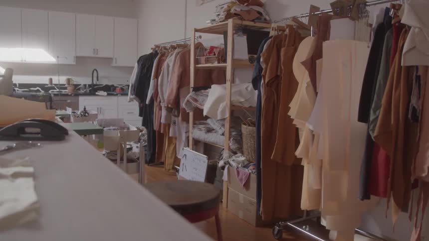 The scene captures a room that appears to be a fashion design studio or workshop with multiple racks of clothing and fabric samples in the background a kitchen area with white cabinets is visible