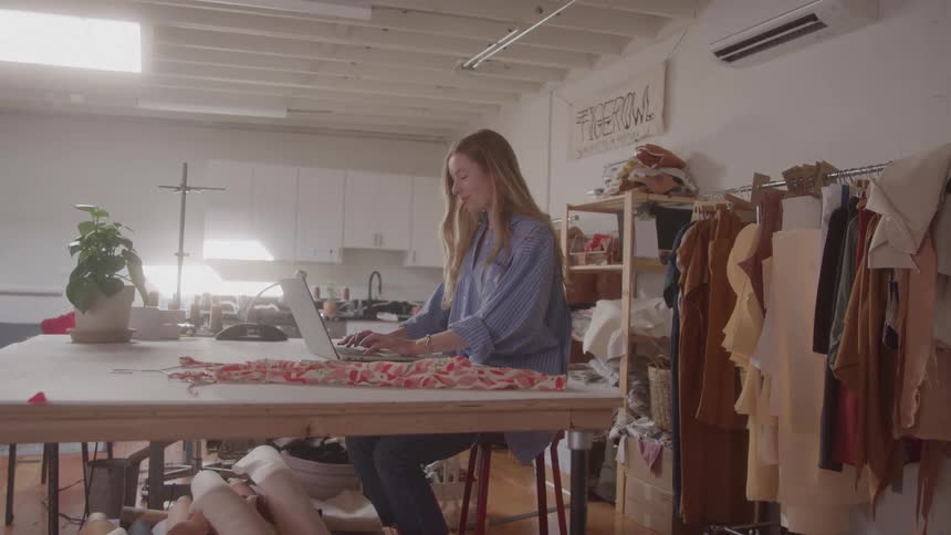 The video shows a woman with long blonde hair sitting at a large work table actively typing on a laptop she is in a creative workspace possibly a fashion studio with racks of clothing and fabric rolls visible around her