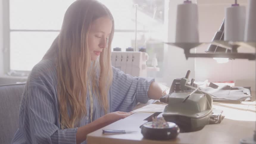 The video shows a woman with long lightcolored hair wearing a striped shirt sitting at a desk and working she appears to be focused on a task involving fabric or paper with a sewing machine and spools of thread visible nearby a filmsupply waterma