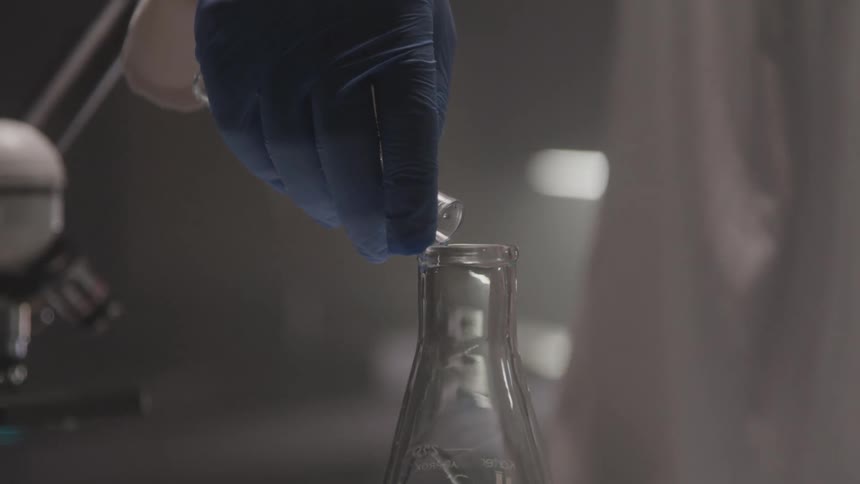 A gloved hand pours blue liquid into a lab flask in a lab