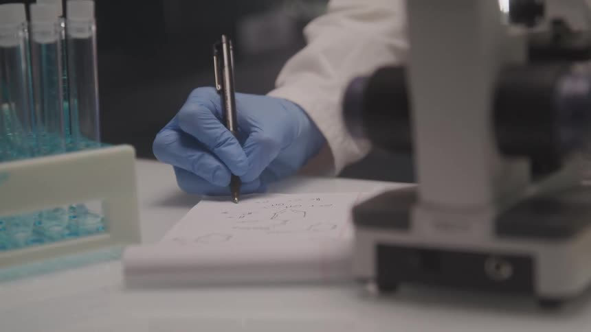 A closeup shot of a lab setting shows a person wearing blue gloves writing on a notepad with a pen next to a microscope and a rack of test tubes with blue liquid the individual appears to be taking notes on what looks like chemical structures