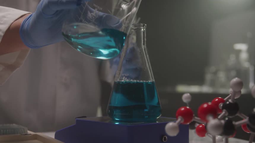 A gloved hand pours blue chemical liquid into a lab flask