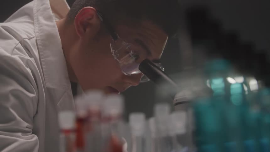 A focused scientist in safety goggles leans over a lab bench surrounded by rows of vividly colored test tubes