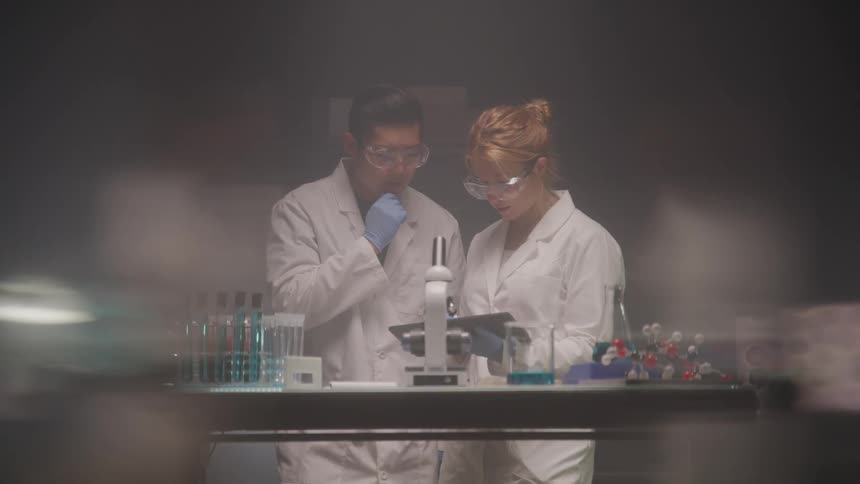 Two lab assistants inside a lab study a folder with a microscope in front of them both wearing safety goggles and white lab coats