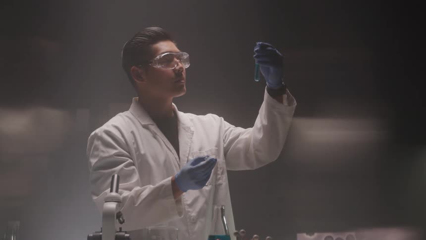 An asian male scientist drops a blue liquid with a pipette into a test tube and inspects it in a lab