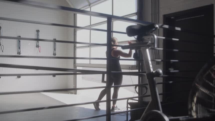 The video shows a person with blonde hair viewed from behind standing in a gym or workout space they are wearing dark athletic wear and appear to be looking out of a large window or glass garage door with gym equipment like a stationary bike visible i