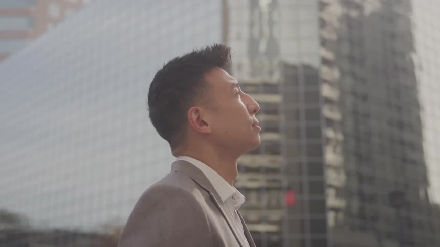 Man in gray blazer looking up while standing in an outdoor setting surrounded by modern city buildings