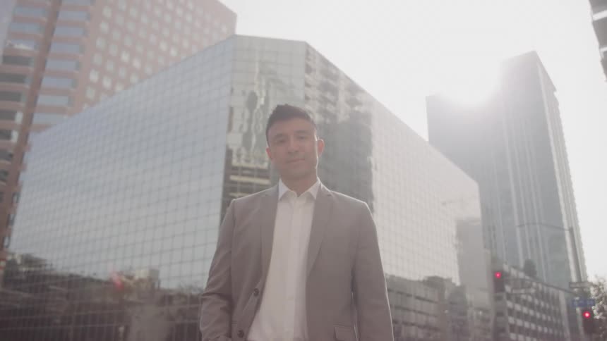 A male model with dark hair and a light beard is standing confidently in front of a modern glass building looking directly at the camera he is wearing a lightcolored suit jacket over a white shirt and the bright sunlight reflects off the building behi