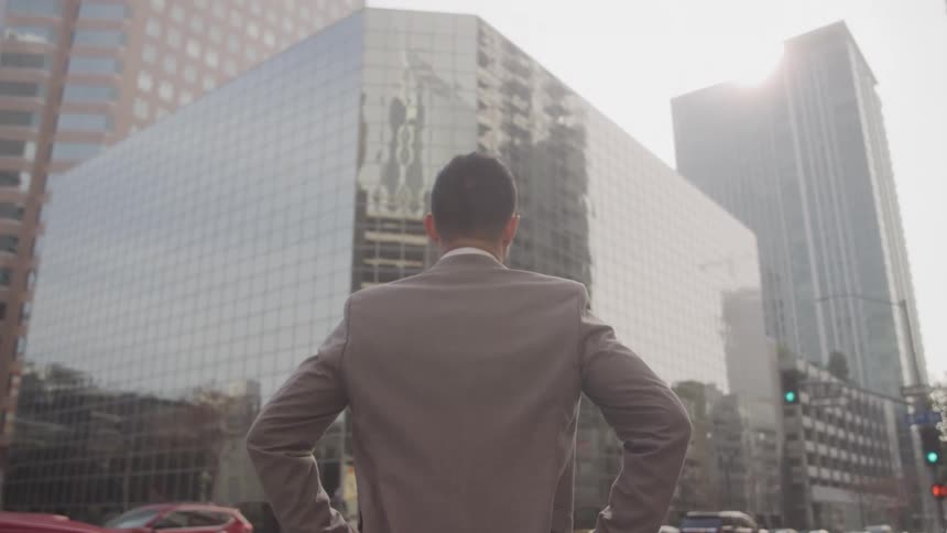 A man stands confidently facing a large glass office building in the city