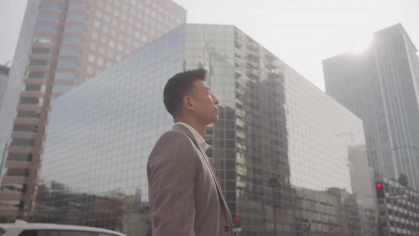 A young man stands in the middle of the street under the sun wearing a suit with a series of highrise and skyscraper buildings in the background