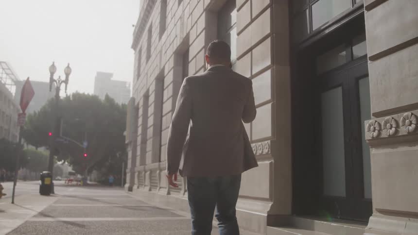 A man in a blazer walks along a quiet city sidewalk while talking on the phone