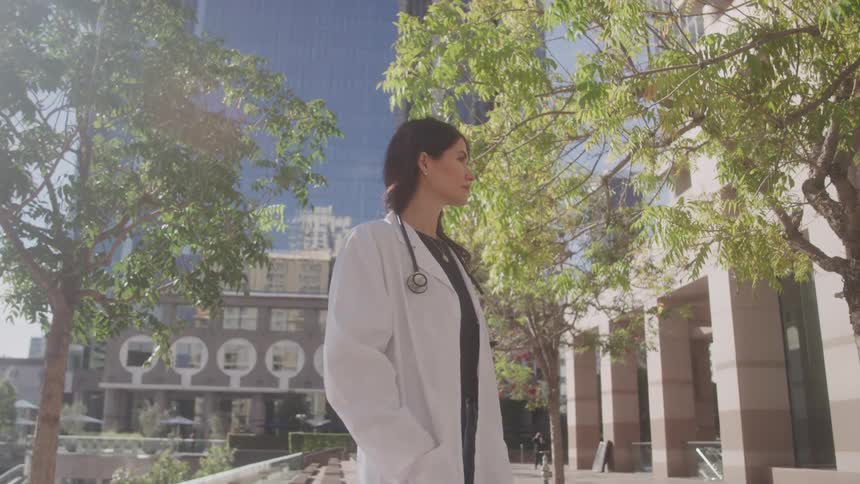 A female doctor wearing a white lab coat and a stethoscope is walking outdoors amidst modern buildings and lush trees the lighting suggests a bright sunny day