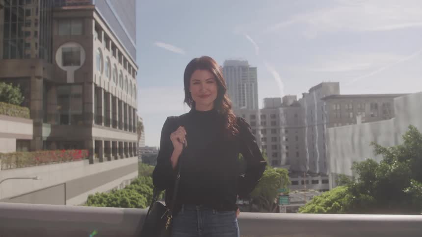 The video features a woman with dark hair wearing a black top and jeans standing outdoors with a city background she is holding a bag over her shoulder and looks towards the camera with a subtle smile a filmsupply watermark is visible in the middle