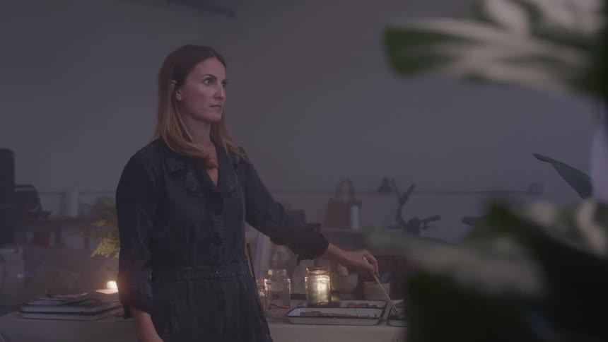 A woman with long straight brown hair wearing a dark jumpsuit stands in a dimly lit room with a contemplative expression she is holding a paintbrush and looking away from the camera with what appears to be a watercolor palette and jars in front