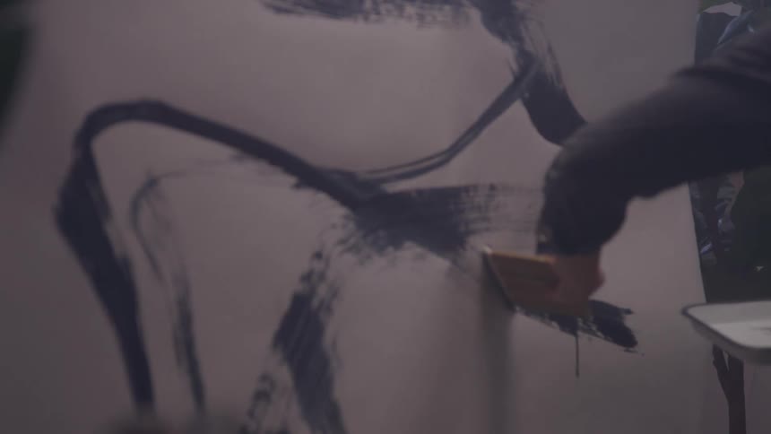 A person paints bold abstract black brushstrokes on a large vertical canvas or wall