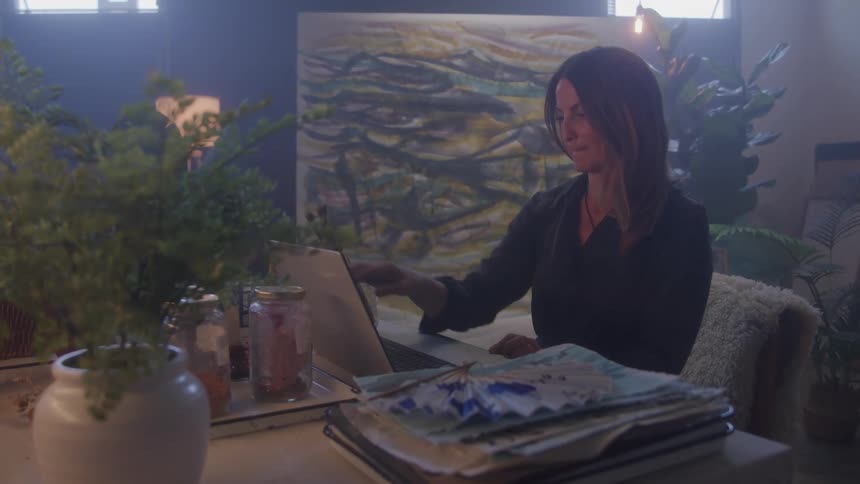 Woman works on laptop in cozy dim room with plants and art supplies