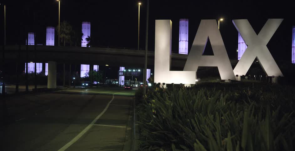 The video captures a night scene at a busy airport entrance with the prominent lax sign illuminated in the foreground cars and buses are seen moving along the road indicating active traffic