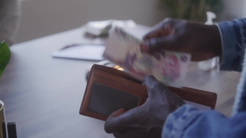 A customer wearing a blue denim jacket takes a bill from a brown wallet and hands it to a cashier across a light colored counter where a plant and payment terminal sit