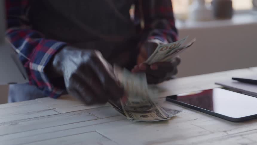 A person is seen from the waist up wearing a plaid shirt counting and sorting several dollar bills on a light colored wooden table with a tablet and papers nearby