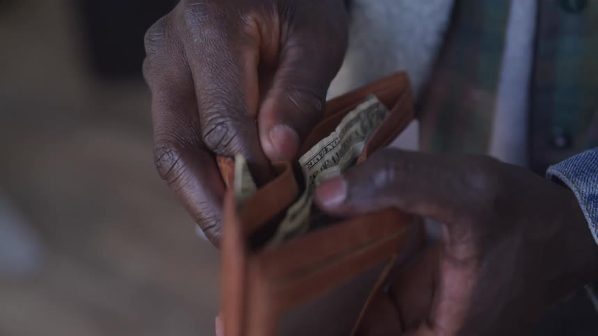A close view shows hands holding a light brown leather wallet with fingers reaching inside to count or retrieve several united states dollar bills