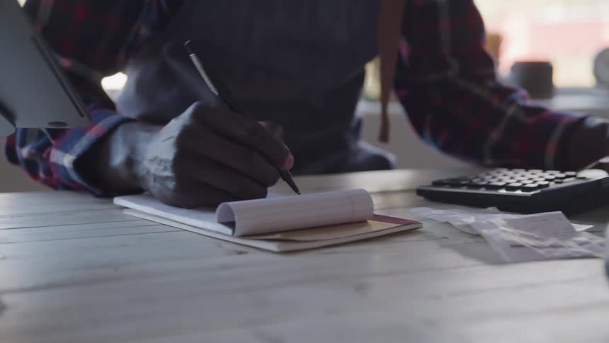 A man at a counter using a calculator to do calculations holding a banknote in one hand then writing something in a notepad