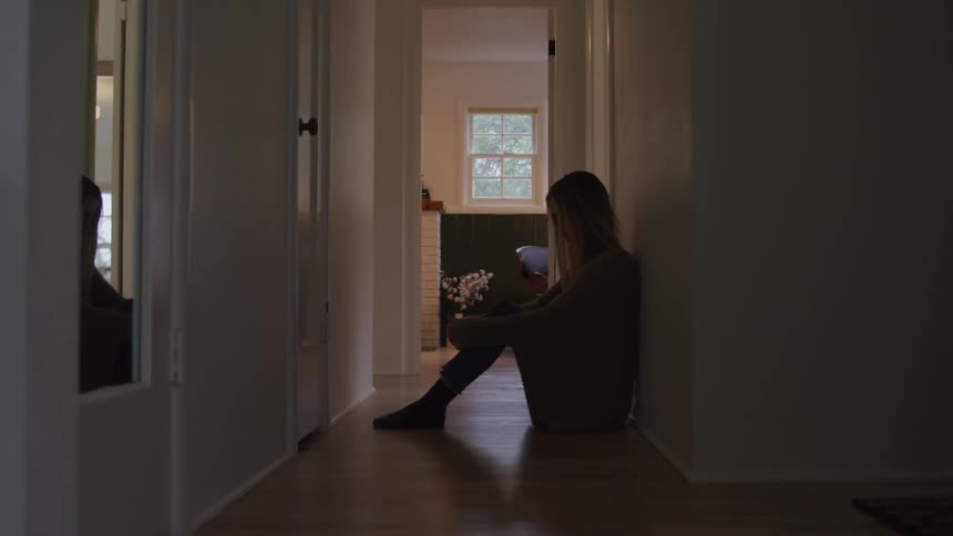 A woman sits on the floor leaning against a wall in a dimly lit hallway looking down with a bright window at the end of the hall