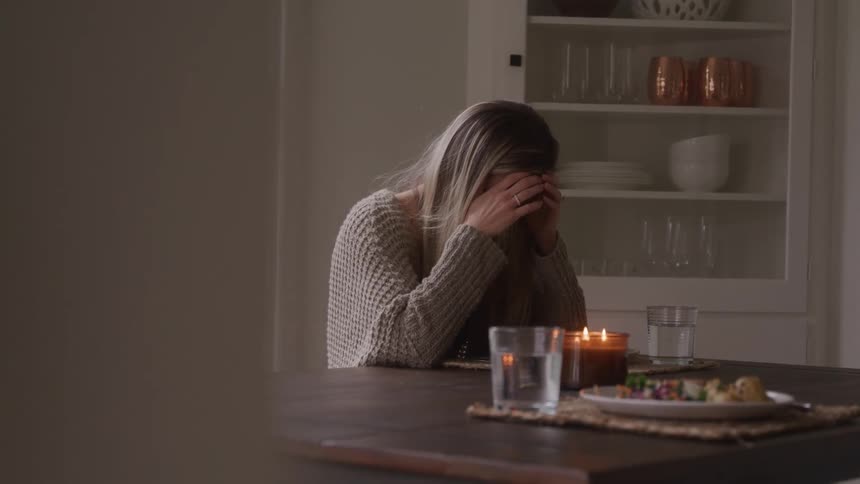 A woman grieves at the dinner table across from a plate of food set for someone else