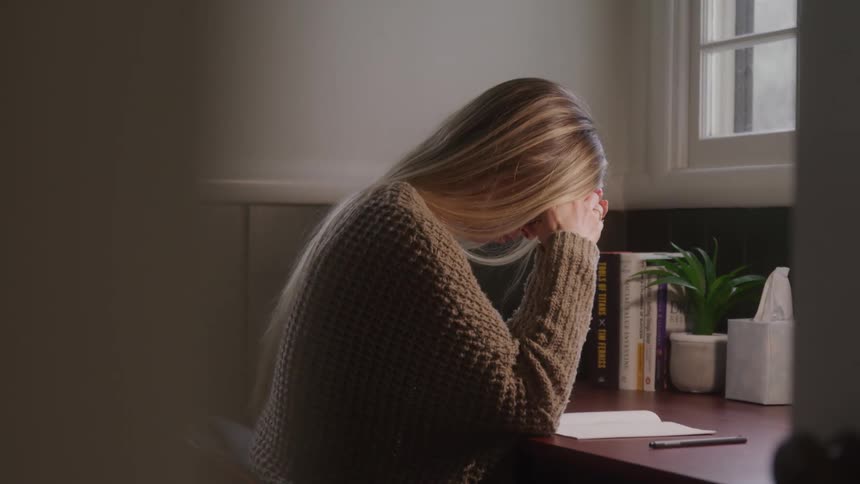 A young woman sits behind a desk indoors at midday reading a book with both hands on her forehead looking tired
