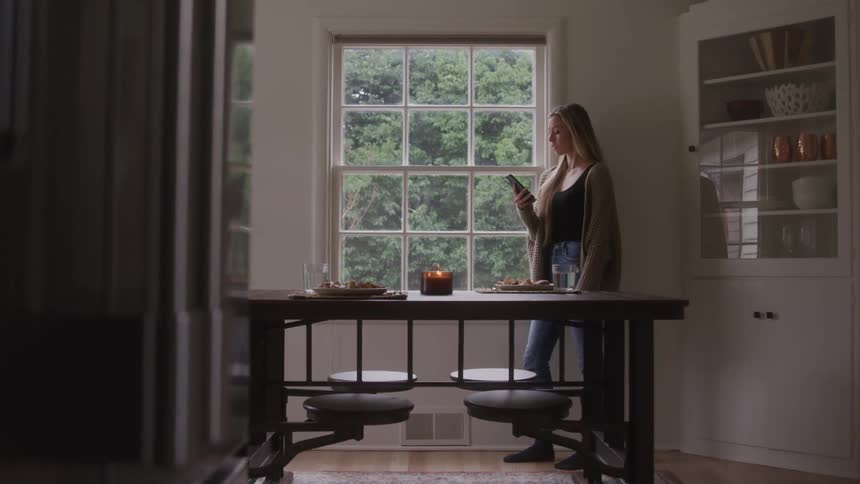 Woman standing by the dining table looking at her phone in front of a window with a view of trees