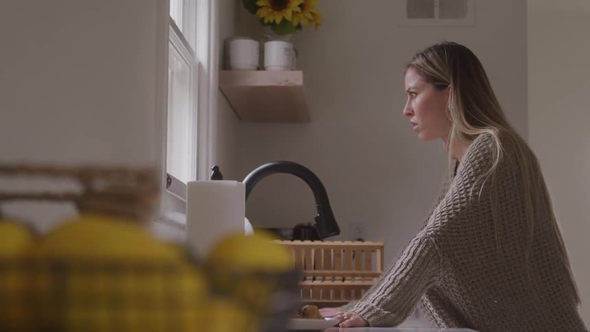 A woman with long blonde hair wearing a knitted sweater leans over a kitchen sink seemingly focused on an activity with food while a basket of yellow fruit is blurred in the foreground