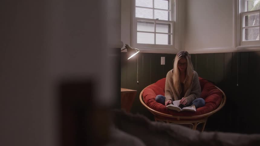 A young woman sits in the corner of her room reading a book with a reading lamp by her side