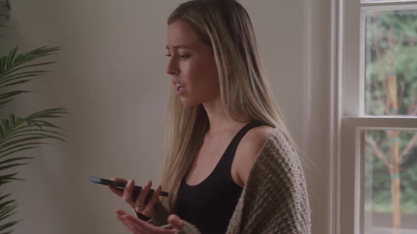 A young adult woman with long blond hair stands by a window holding a smartphone and gesturing with her free hand talking on the phone