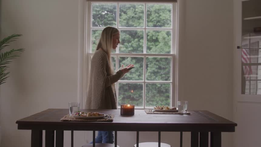 The woman paces by the dining table while talking on the phone