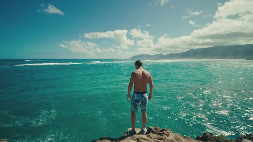 A young man wearing swimming trunks stands on a cliff ready to jump into the ocean on a sunny day
