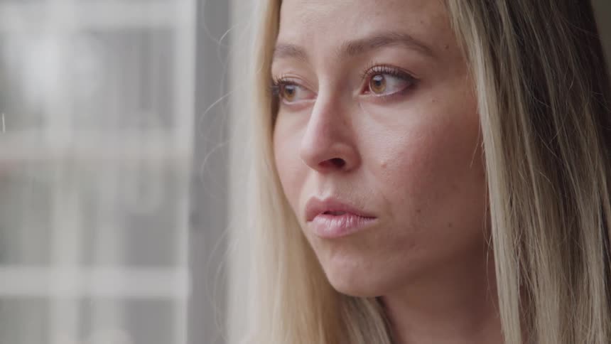 A young adult woman with long blond hair looks out a window with a pensive expression her gaze directed towards the right side of the frame