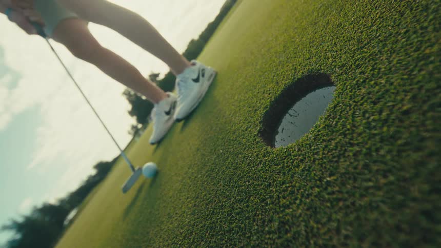 The video shot from a low angle on a golf green shows a persons feet in white golf shoes poised over a golf ball next to the hole the person then putts the ball which goes into the hole and they reach down to retrieve it
