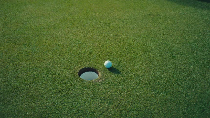 A person plays golf as the ball rolls down the course close to the hole