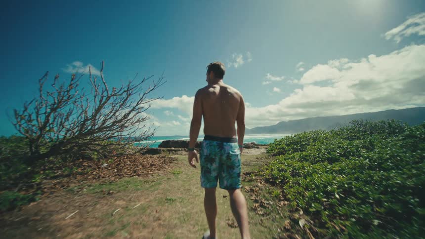 A man with no shirt on and wearing patterned shorts walks away from the camera on a dirt path through low green bushes towards a bright blue ocean with rolling waves under a clear sky with scattered clouds