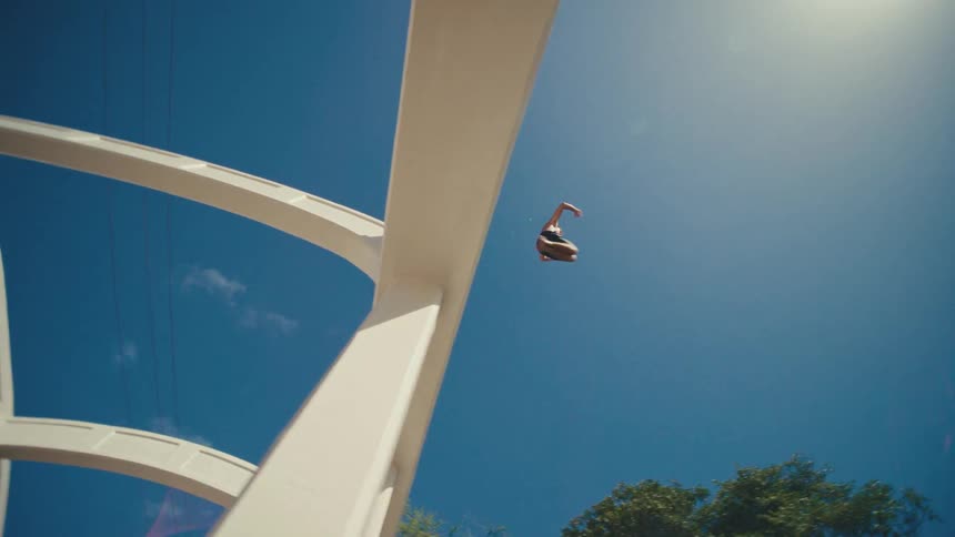 A young man jumps off a high bridge into a river at midday creating a splash on a sunny day then swims to shore