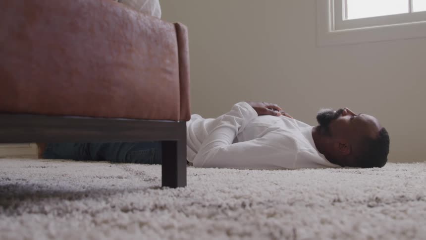 A middleaged man lies on a rug on the floor next to his bed indoors at midday
