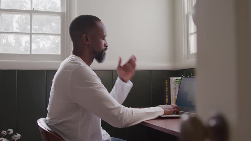 A man sits behind a desk at midday working on a laptop and looking sad