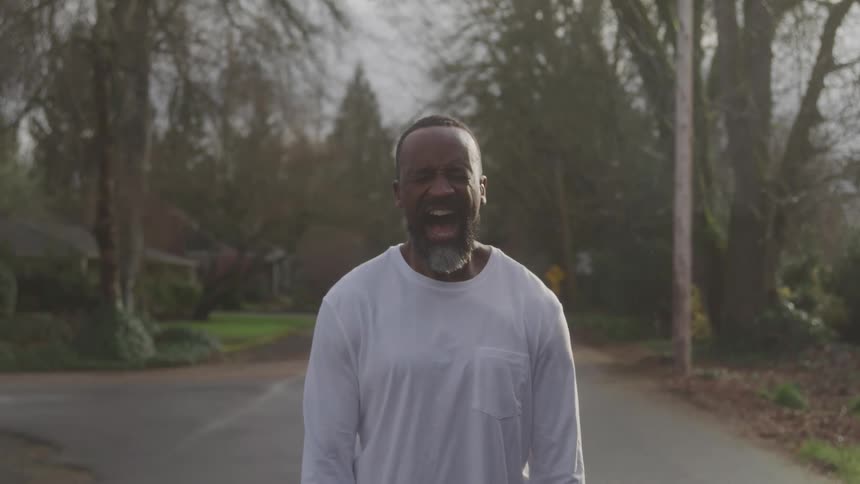 An older adult man with a beard and short hair wearing a white long sleeved shirt stands on a paved road looking directly at the camera screaming or shouting with trees and residential properties in the background