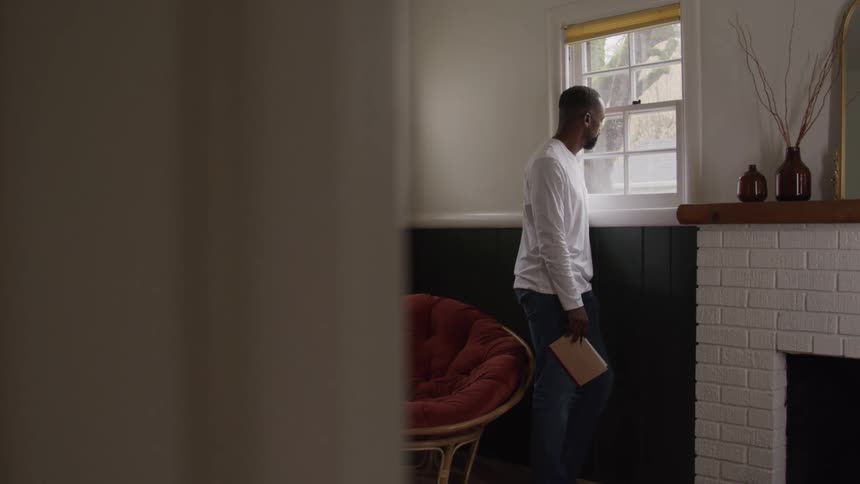 A man stands near a window quietly holding a book as he looks outside with a contemplative gaze in the soft morning light