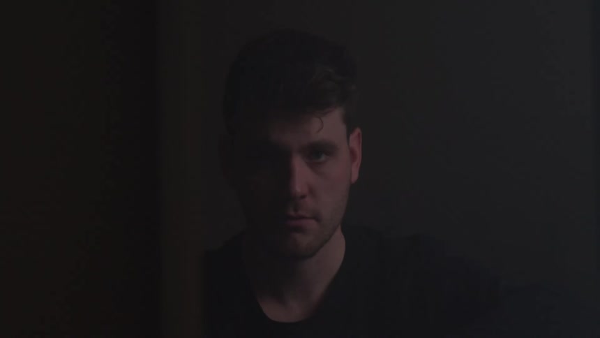 Man stares directly with a serious expression in a dimly lit and softly illuminated setting