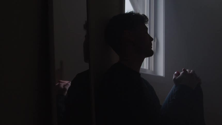 A young man sits inside a room looking sad