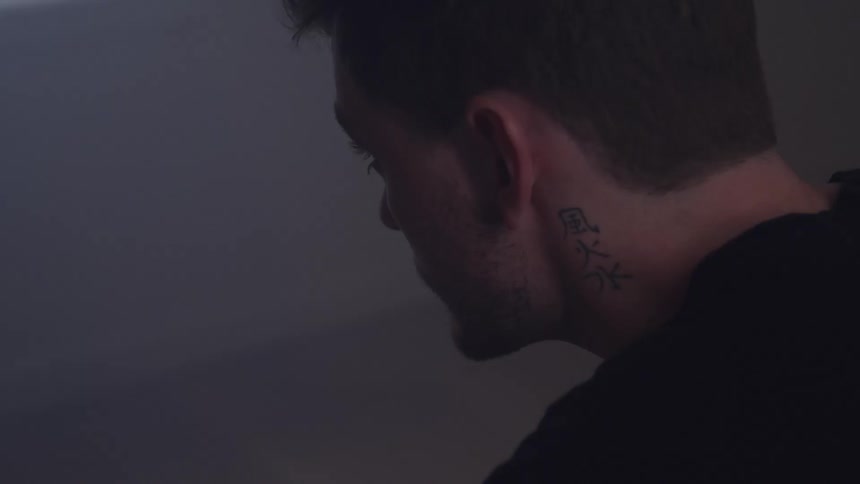 A man with short hair and a neck tattoo looks downward in a dimly lit space his face turned away