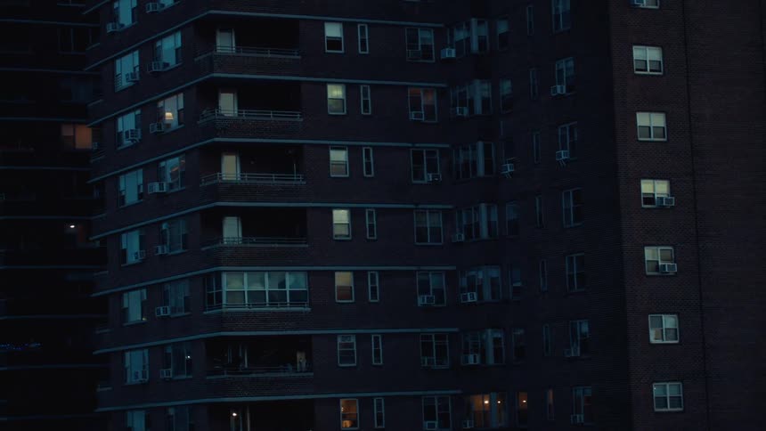 A dark densely packed apartment building shows scattered lights glowing from various windows as evening sets in