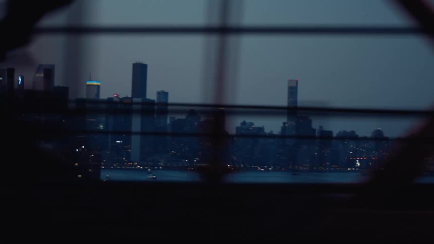 A view of a city skyline at dusk or night partially obscured by bridge girders with distant lights twinkling and a body of water in the foreground