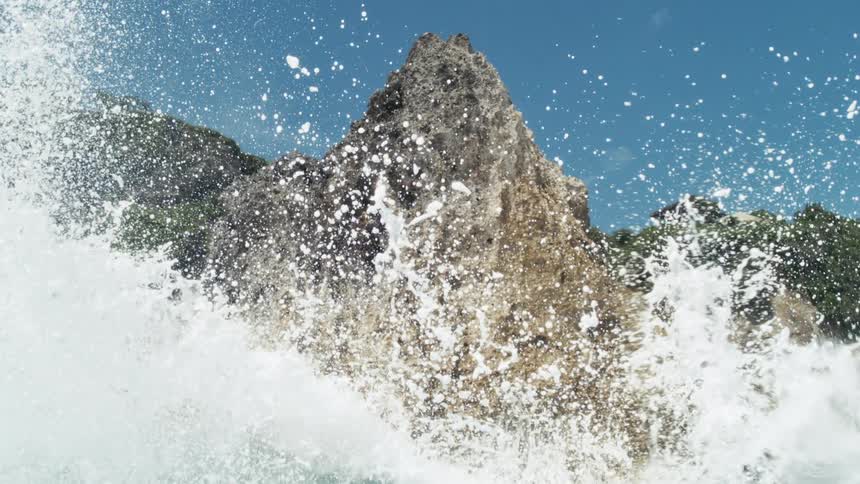 Ocean waves crash violently against a jagged coastal rock formation beneath a clear blue sky