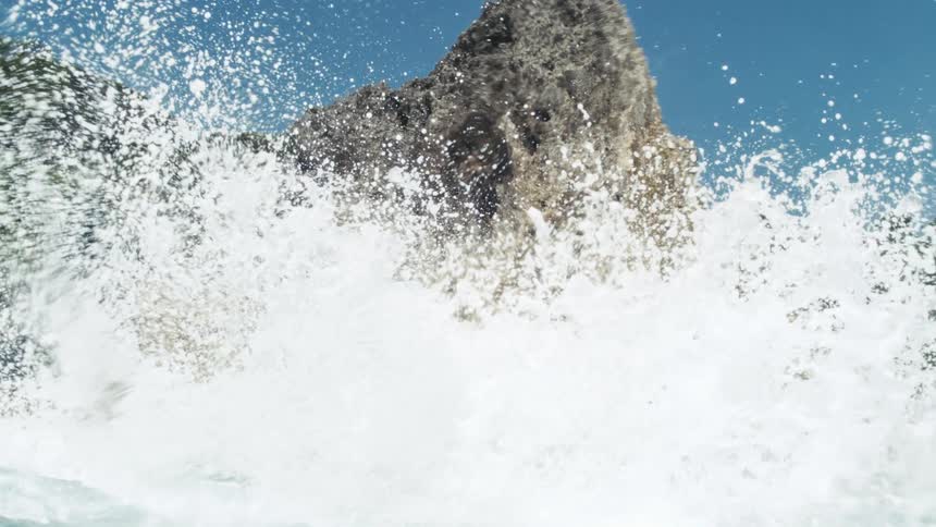 Waves crash against a large textured rock formation on a sunny day sending white spray and droplets into the clear blue sky